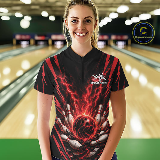 A woman wearing a black quarter zip bowling jersey with a red and black lightning graphic and bowling pins holding a bowling ball, standing in a bowling alley.