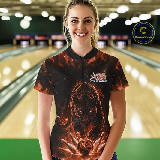 Front view of a female model in a bowling lane wearing a black quarter-zip shirt with glowing electric panther design and bowling ball striking pins in orange and black style.