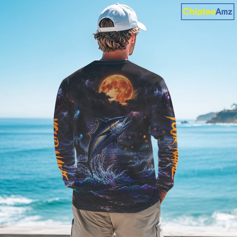 A man stands with his back to the camera wearing a long-sleeve graphic shirt featuring a large jumping fish and a glowing full moon on a dark background, with orange text down the sleeves, set against an ocean backdrop.