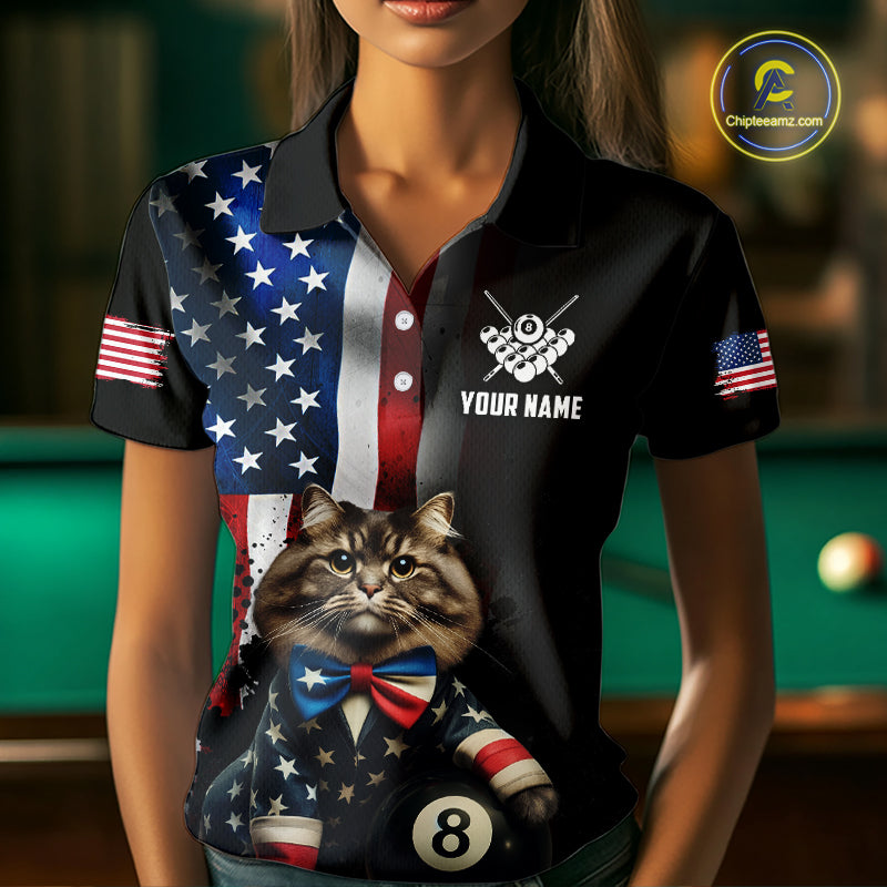 A front view of a custom black billiards polo shirt worn by a female model. The shirt features a distressed US flag design on one side and a cartoon cat in a vest with its paw on an 8-ball on the front. A 'YOUR NAME' text placeholder is positioned below a crossed pool cues icon. The background is a blurred billiards room with a green pool table.