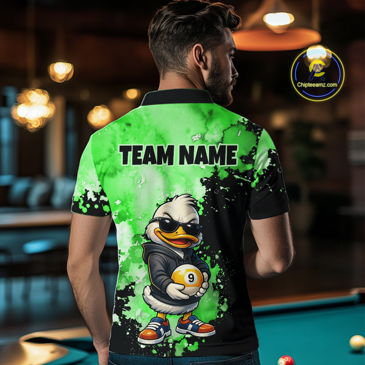 A man viewed from behind wearing a custom-printed green and black polo shirt with a cartoon duck graphic and the text 'TEAM NAME' on the back. The shirt features a vibrant green and black watercolor splatter design with a cartoon duck character holding a billiard ball.