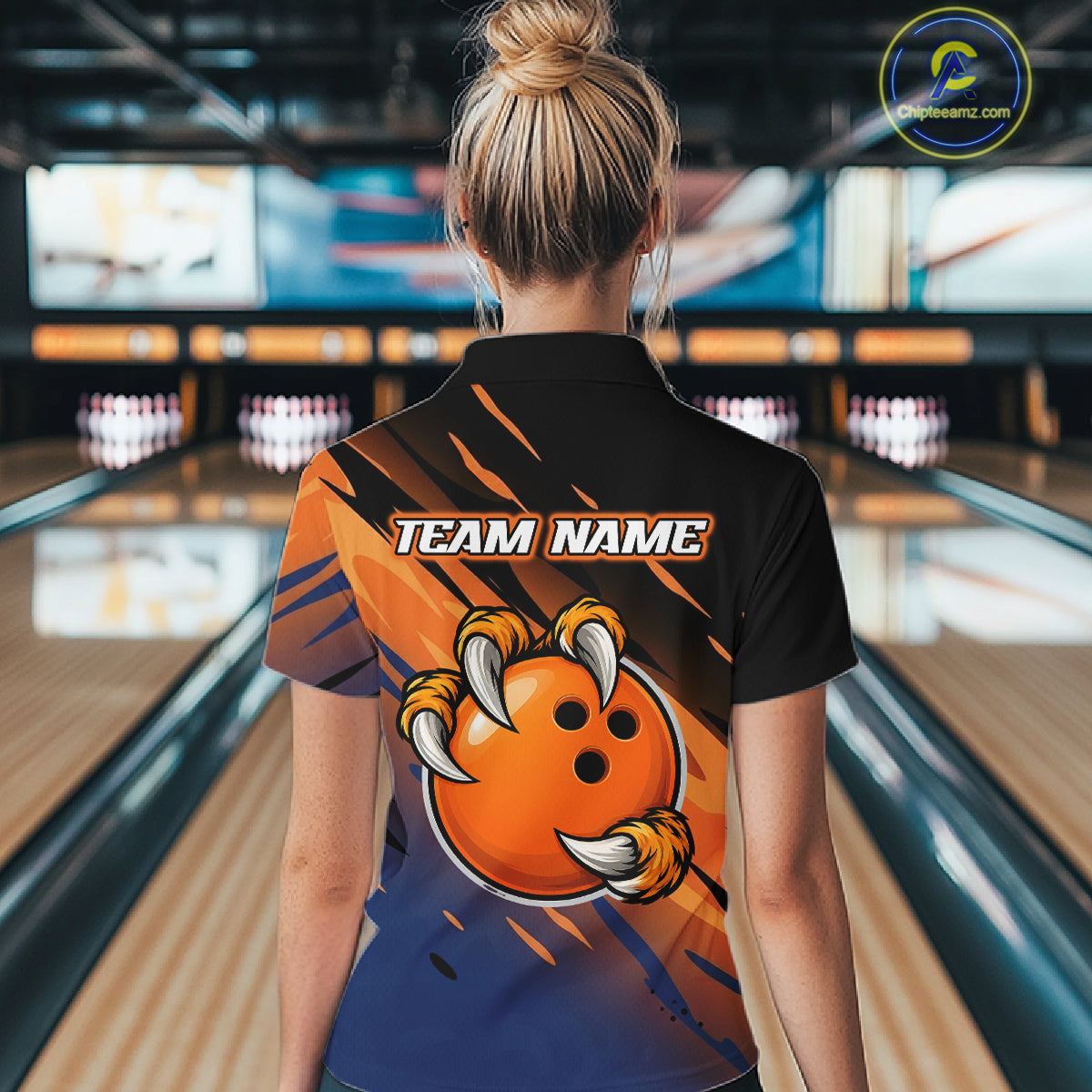 A woman viewed from behind wearing a custom bowling shirt with a clawed orange bowling ball graphic and 'TEAM NAME' text, standing in a bowling alley.