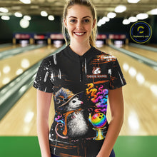 Load image into Gallery viewer, A woman wearing a black short-sleeve bowling-themed t-shirt with a colorful cartoon opossum, bowling ball, and paint splatter design, standing in front of a blurred bowling alley background. 