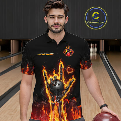 A man wearing a black short-sleeve polo shirt with a fiery flame-bomb bowling graphic, standing in a bowling alley. The shirt features orange and yellow flames, a black bowling ball and claw hand, and the text 'YOUR NAME' on the chest.
