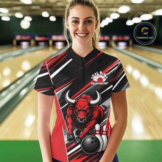 A woman stands in a bowling alley wearing a short-sleeve bowling quarter zip jersey with a red bull graphic, black and red diagonal stripes, and a black collar. 