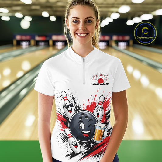 A woman stands in a bowling alley wearing a white short-sleeve quarter-zip bowling shirt with a cartoon bowling ball and pins graphic on the front. The background shows blurred bowling lanes and pins.