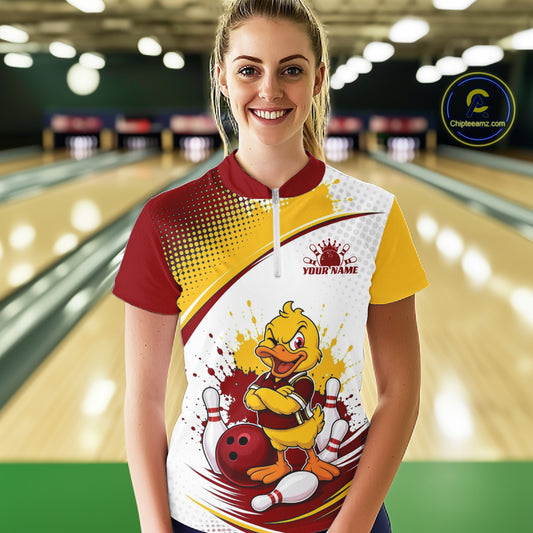 A woman stands in a bowling alley wearing a custom short-sleeve quarter zip bowling shirt with a yellow and red duck mascot graphic on the front. The shirt features a white, yellow, and red color-blocked design with a halftone pattern on the left side and a white front panel with a cartoon duck holding bowling pins and a bowling ball.