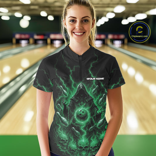 A woman wearing a black bowling shirt with a green and black grim reaper bowling graphic design, standing in front of a blurred bowling alley background. 