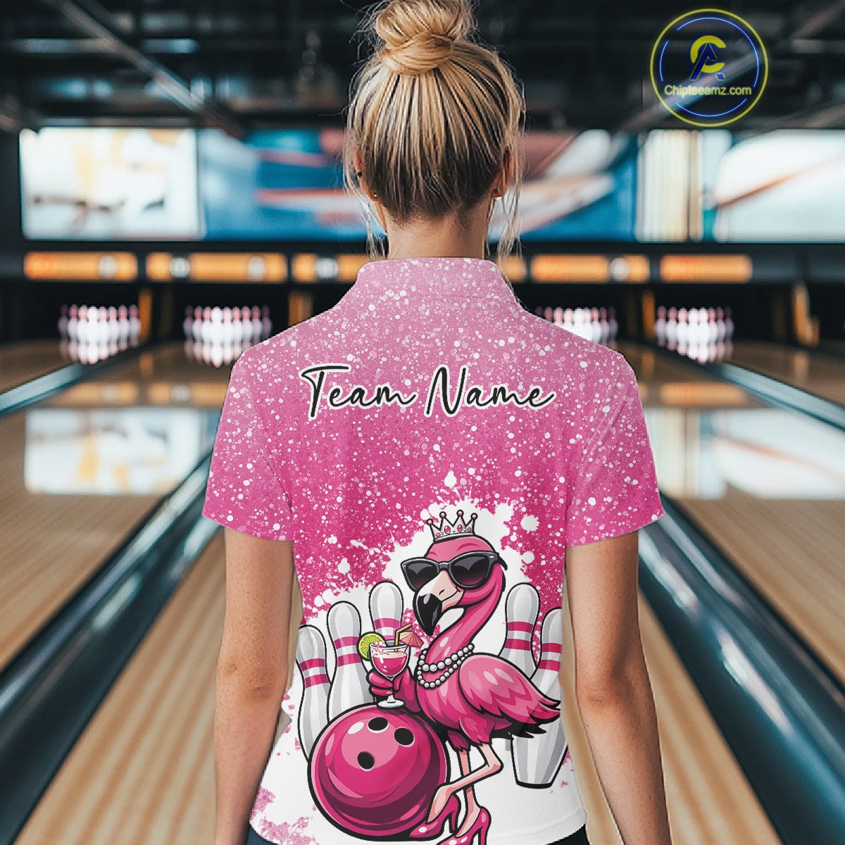 Back view of a female model in a bowling alley wearing a pink polo shirt featuring a crowned flamingo holding a cocktail beside a bowling ball.