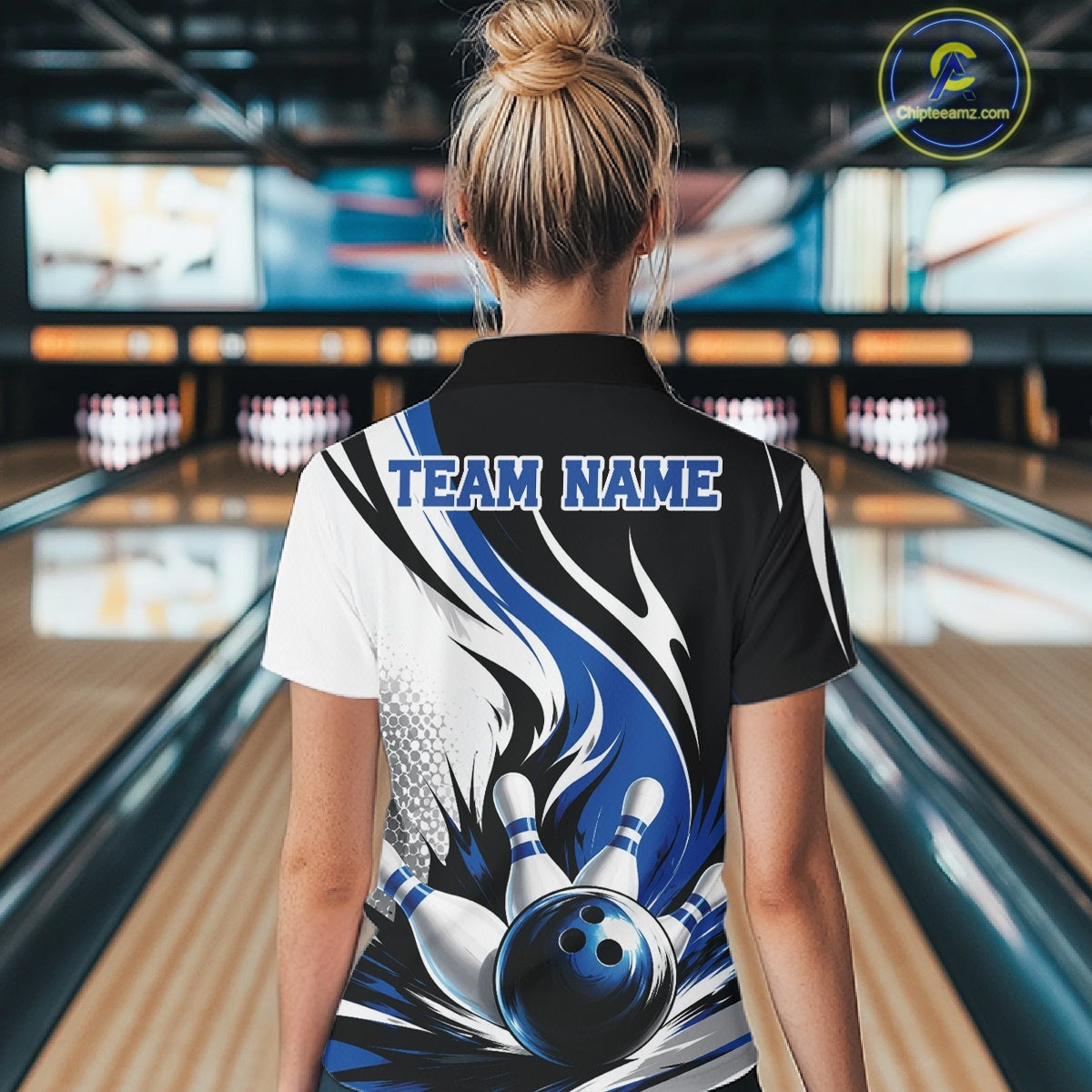 Back view of a female model wearing a white, black, and blue bowling quarter zip shirt with a flaming bowling ball knocking down pins graphic at a bowling lane.