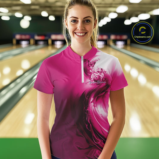 Female model wearing a pink bowling quarter zip with butterfly graphics and bowling artwork, front view at the bowling lane