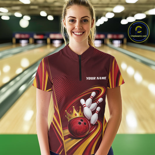 A woman standing in a bowling alley wearing a custom short-sleeve bowling quarter zip shirt with a maroon, yellow, and red graphic design featuring bowling pins, a bowling ball, and a crown. 
