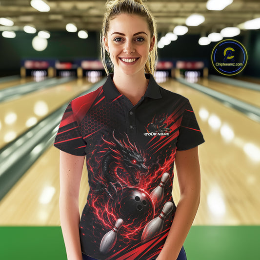 A woman stands in a bowling alley wearing a black short-sleeve polo shirt with a red and black dragon graphic wrapping around the front, with bowling pins and balls depicted.