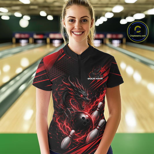 A woman wearing a black and red bowling-themed short-sleeve quarter zip shirt with a dragon and bowling ball graphic, standing in a bowling alley.