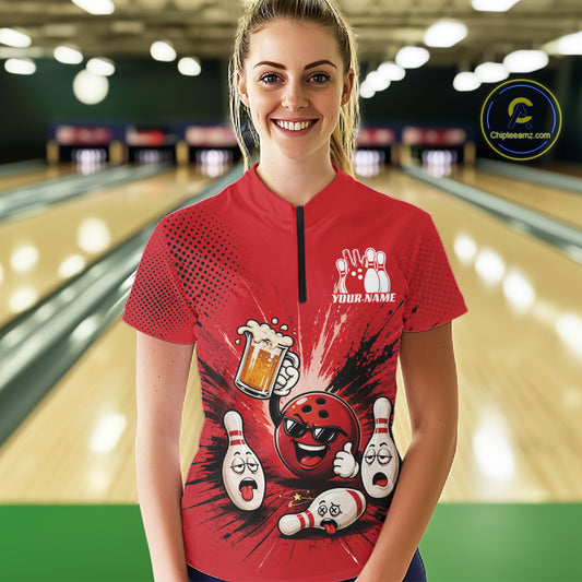 A woman stands in a bowling alley wearing a red short-sleeve quarter-zip bowling shirt with a cartoon bowling ball and pins graphic on the front. The background shows blurred bowling lanes.