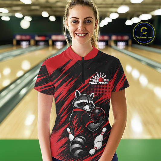 A woman stands in a bowling alley wearing a bowling short-sleeve quarter zip shirt with a red and black brushstroke design, a raccoon graphic holding bowling pins