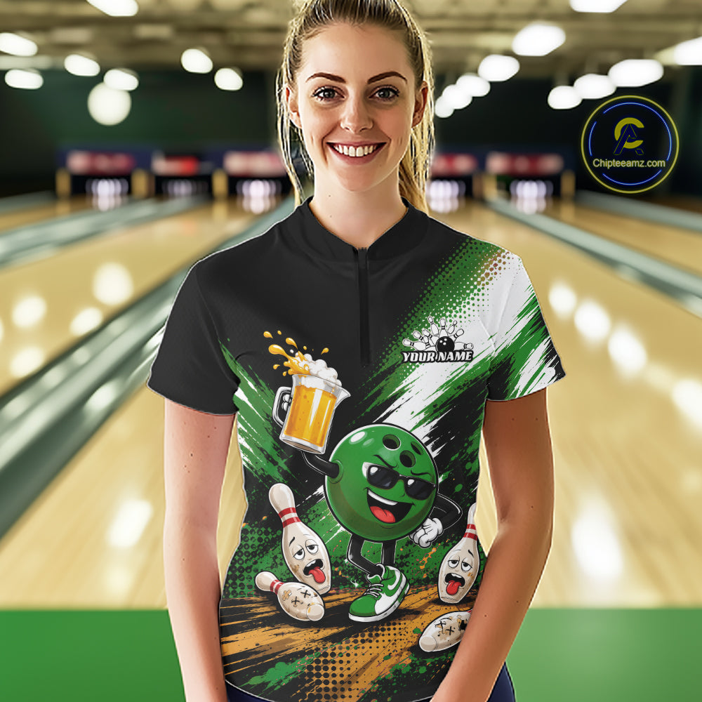 A woman stands in a bowling alley wearing a custom short-sleeve quarter zip shirt with a green and black bowling-themed graphic print featuring a funny cartoon bowling ball with a beer pitcher and pins. 
