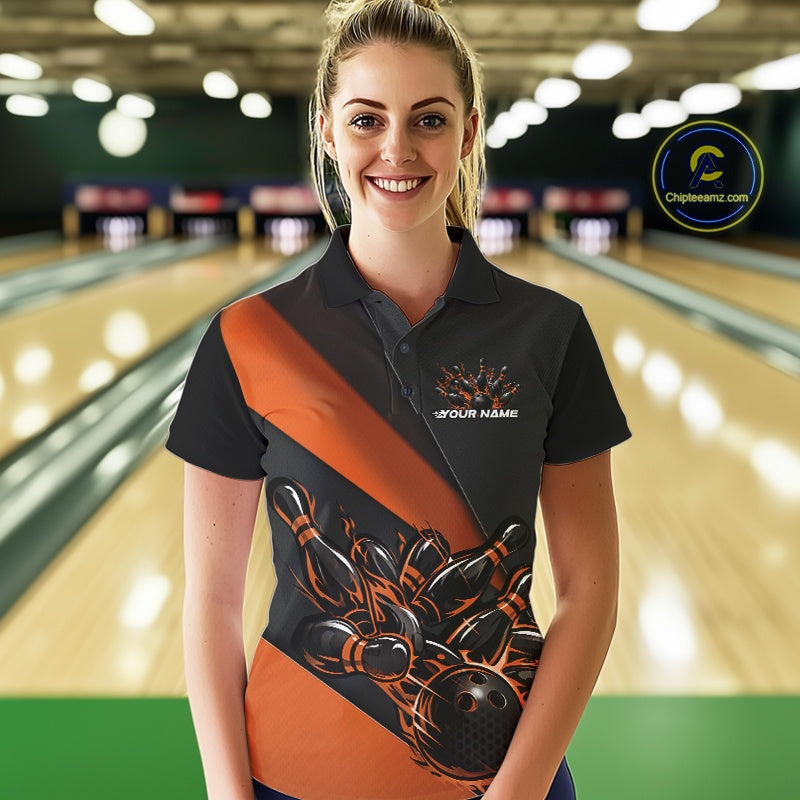 Female model front view in a bowling lane wearing a black and orange polo shirt featuring flame bowling ball striking pins in dynamic sports artwork.