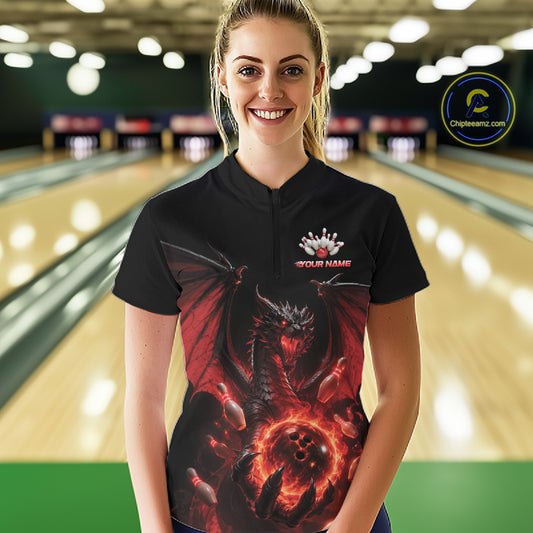 A woman standing in a bowling alley wearing a black short-sleeve bowling quarter zip shirt with a red dragon graphic and flaming bowling ball