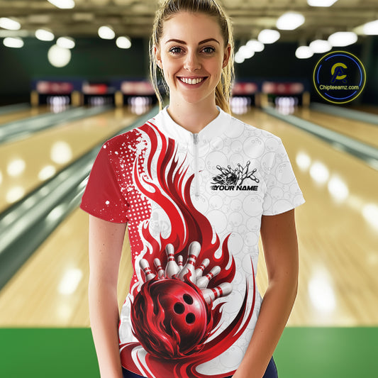 A woman stands in a bowling alley wearing a custom short-sleeve bowling shirt with a red and white flame-and-bowling-ball graphic. The shirt has a white collar and a black placeholder logo on the left chest that reads 'YOUR NAME'.