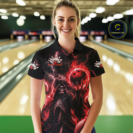 A woman wearing a black short-sleeve polo shirt with a red and black horror-themed graphic print featuring a skeleton and a bowling ball, standing in a bowling alley.