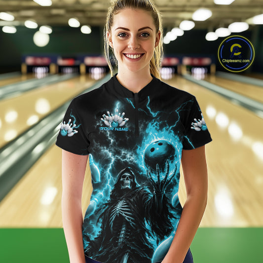 A woman stands in a bowling alley wearing a black short-sleeve quarter-zip shirt with a dramatic blue and black graphic print featuring lightning, a skull, and a bowling ball. 