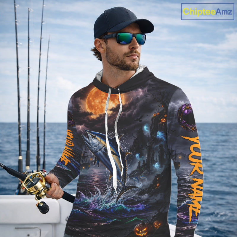 A man wearing a black hoodie with a detailed all-over Halloween fishing graphic print stands on a boat deck. The hoodie features a large blue fish jumping from the water under a glowing orange full moon, with jack-o'-lanterns and orange vertical text on the sleeves.