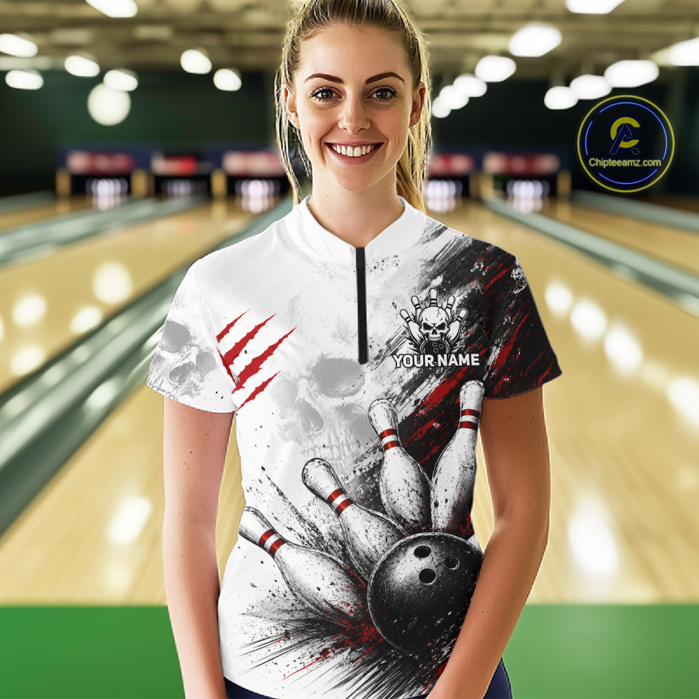 A woman stands in a bowling alley wearing a custom short-sleeve bowling shirt with a white, black, and red graphic design featuring bowling pins, a bowling ball, claw marks, and a skull. The shirt has a mock neck with a black zipper and the text 'YOUR NAME' on the chest.
