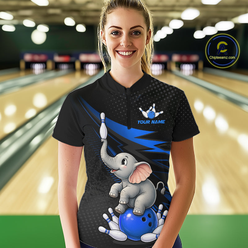 A woman stands in a bowling alley wearing a black short-sleeve bowling shirt with a cartoon baby elephant graphic on the front, blue lightning accents, and a blue bowling pin logo. The background shows blurred bowling lanes.