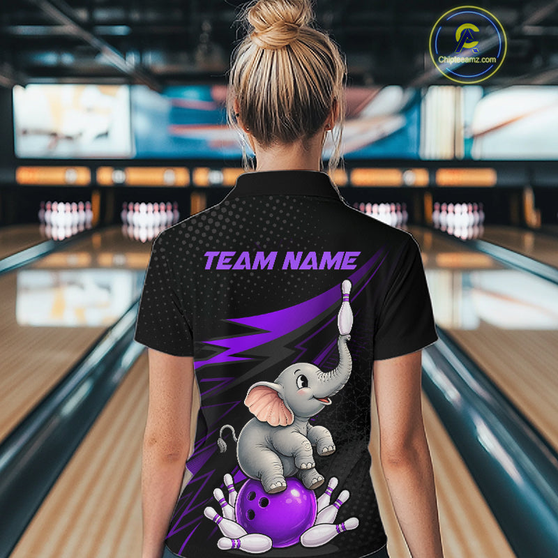 A woman viewed from behind wearing a black bowling-themed polo shirt with a cartoon baby elephant on a purple bowling ball, purple lightning accents, and the text 'TEAM NAME' on the back. The background shows a blurred bowling alley with lanes and pins.