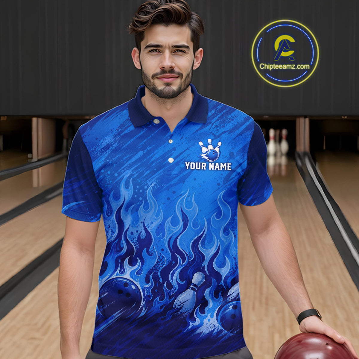 A man wearing a blue short-sleeve polo shirt with a flame and bowling ball graphic, standing in a bowling alley. The shirt has a white crown logo and the text 'YOUR NAME' on the chest.