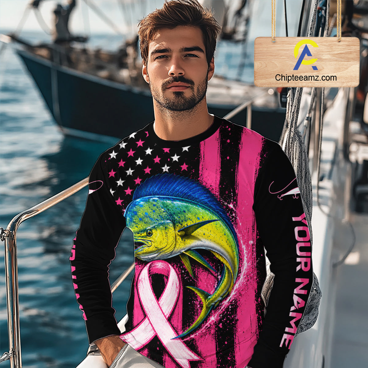 A man wearing a long-sleeve custom graphic shirt with a colorful fish design, pink and black stripes, and a pink ribbon, photographed outdoors near boats with a visible 'Chipleeamz