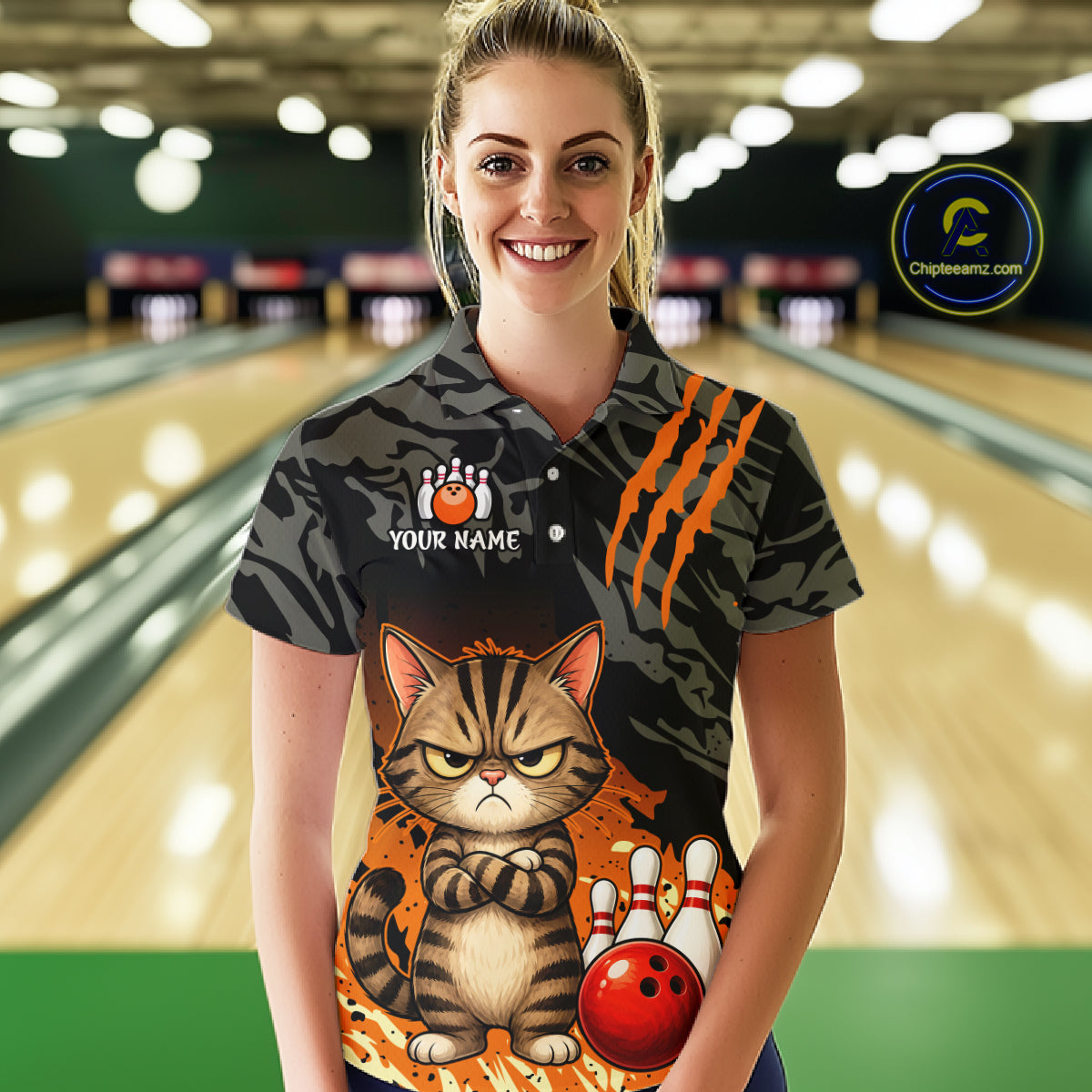 A woman wearing a custom short-sleeve polo shirt with a grumpy cat bowling design, standing in front of a blurred bowling alley background. The shirt features a dark gray and black tiger-stripe pattern with orange claw marks, a large cartoon cat graphic, bowling pins, and a red bowling ball, with a customizable name patch on the chest.