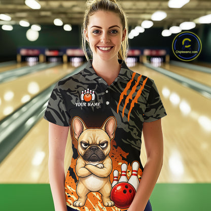 A woman stands in a bowling alley wearing a short-sleeve polo shirt with a dark camouflage pattern, orange claw-stripe graphics, a cartoon French bulldog, bowling pins, and a red bowling ball. The shirt also has a customizable name patch on the chest.