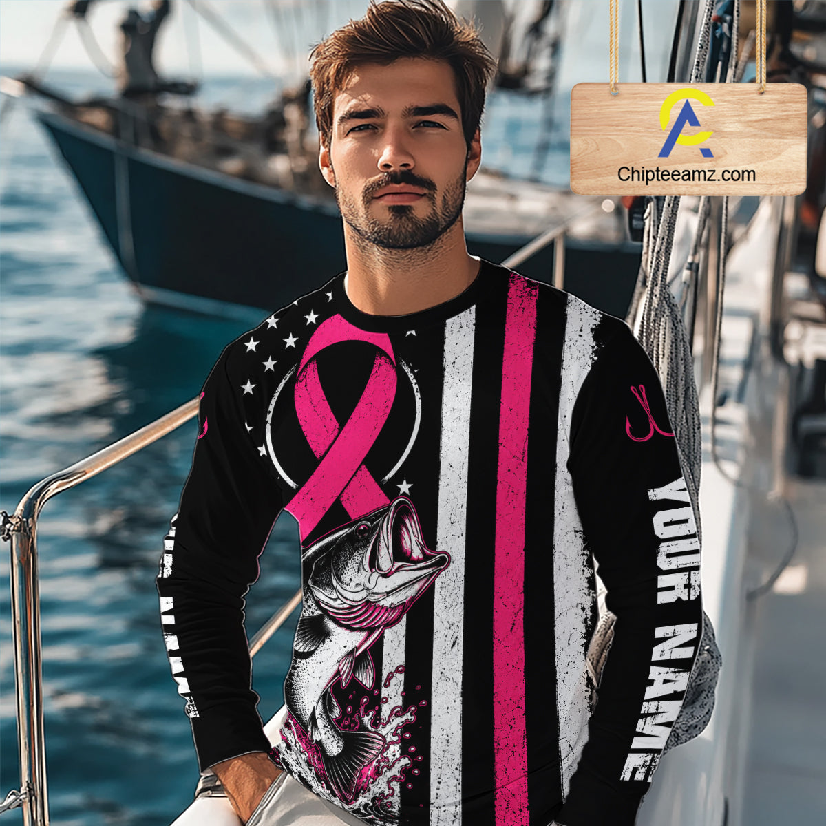 A man wearing a long-sleeve black and white distressed graphic shirt with a pink ribbon, a fish illustration, and 'YOUR NAME' printed on the sleeve, standing on a boat with water and another boat in the background.